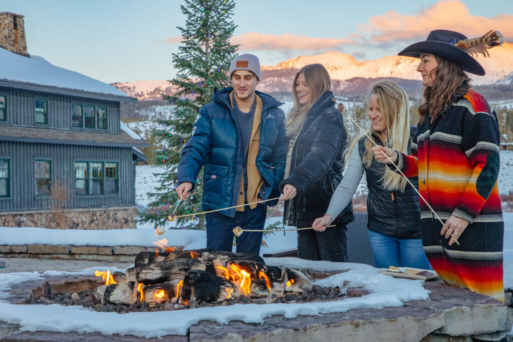 A group of 3 girls and 1 boy are toasting marshmallows over an open fire outside of a resort in the winter. 