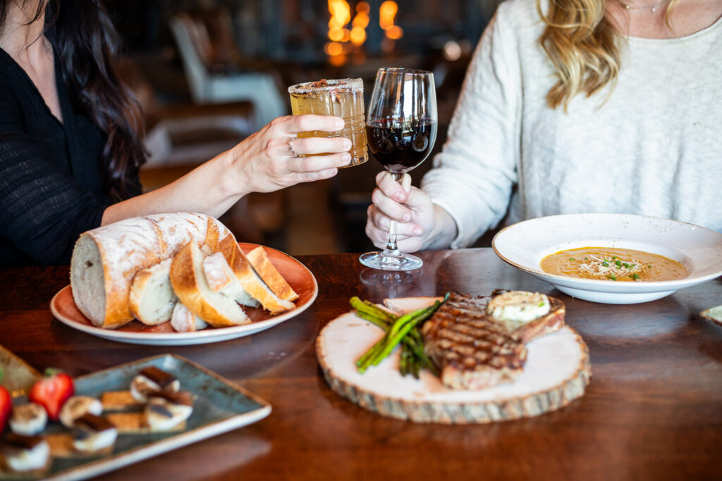 Two people clink their drinks, a red wine and a cocktail, sitting at a table with rustic bread, a steak, soup, and s'mores.