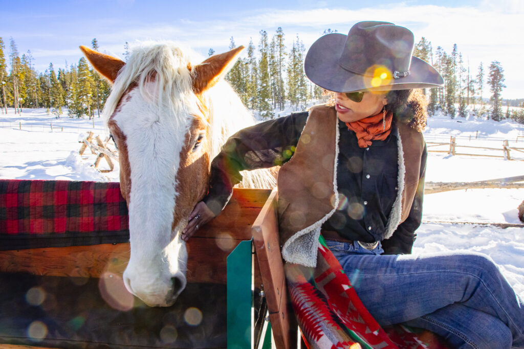 A cowgirl is sitting on the back of a sleigh and petting a horse.