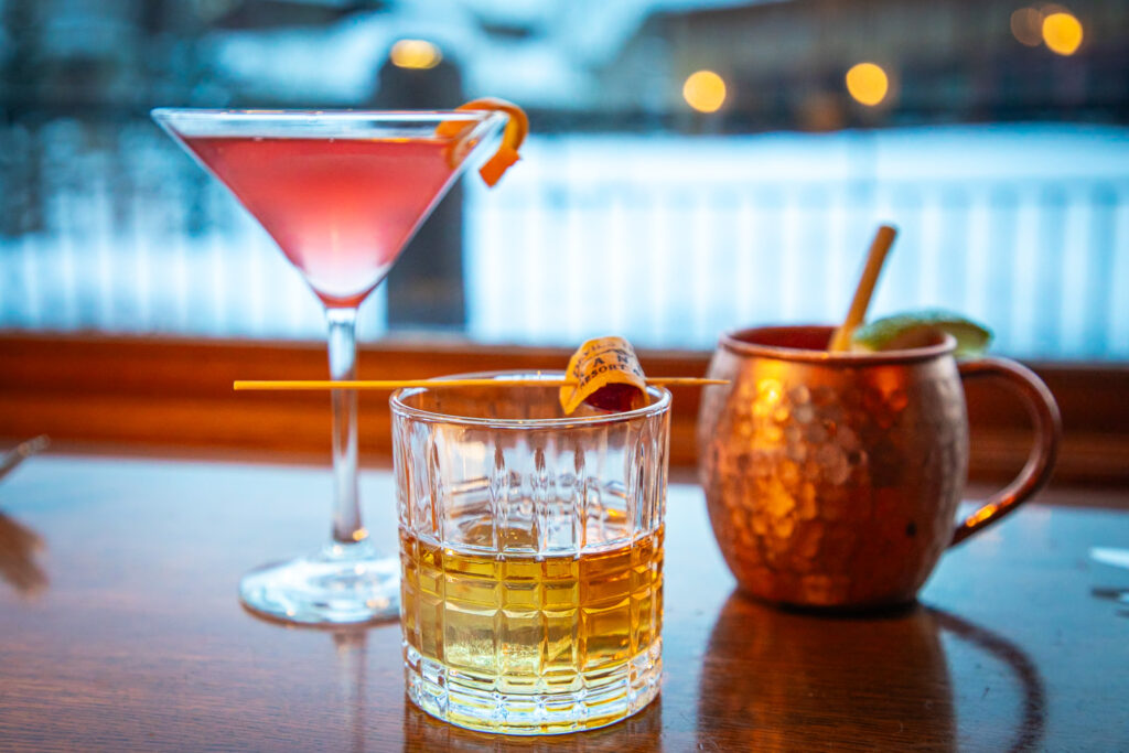 A pink cocktail in a martini glass, a cocktail in a whiskey glass, and a cocktail in a copper mug sit on a table.