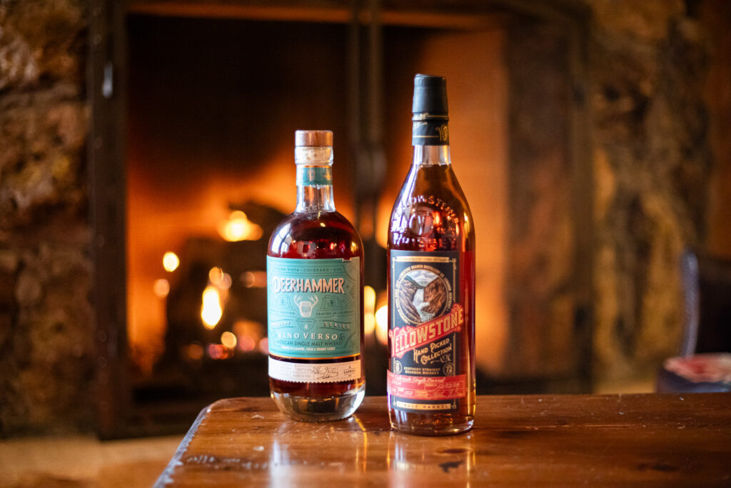 Two whiskey bottles sit on a table with a glowing fireplace in the background.