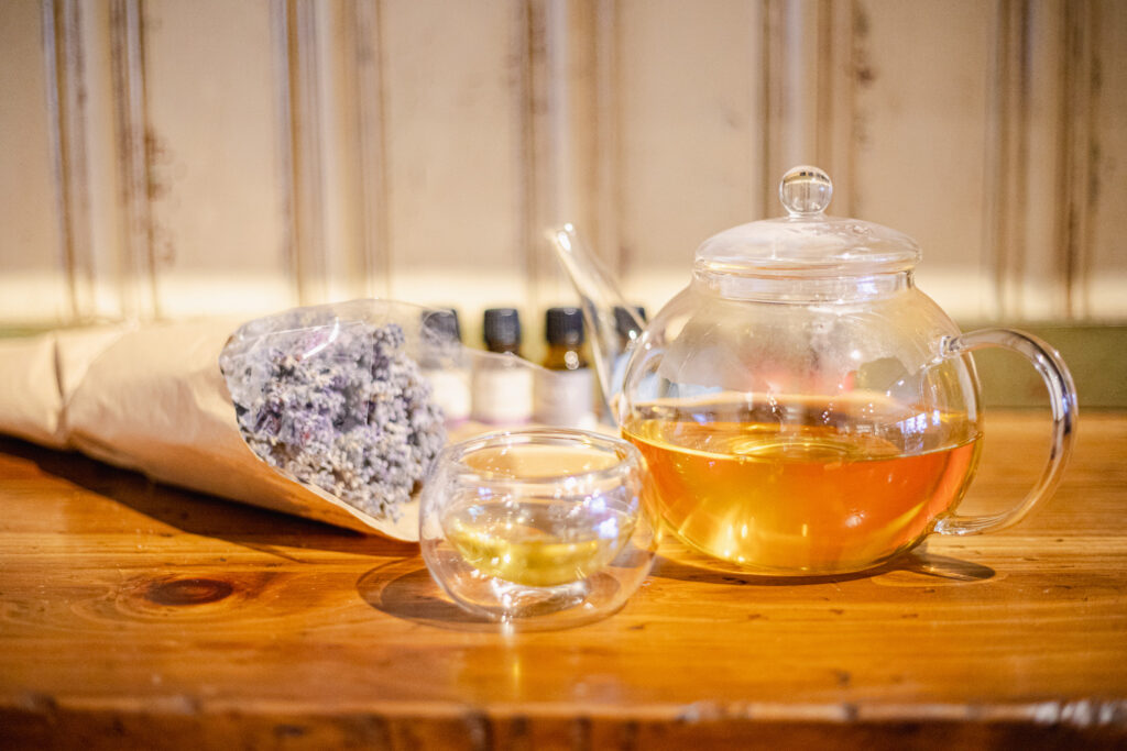 A teapot and teacup with tea sit on a wooden table with a lavender bunch and essential oils in the background.