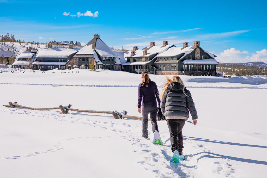 Snowy landscape with mountain lodge and two snowshoers walking in the snow.