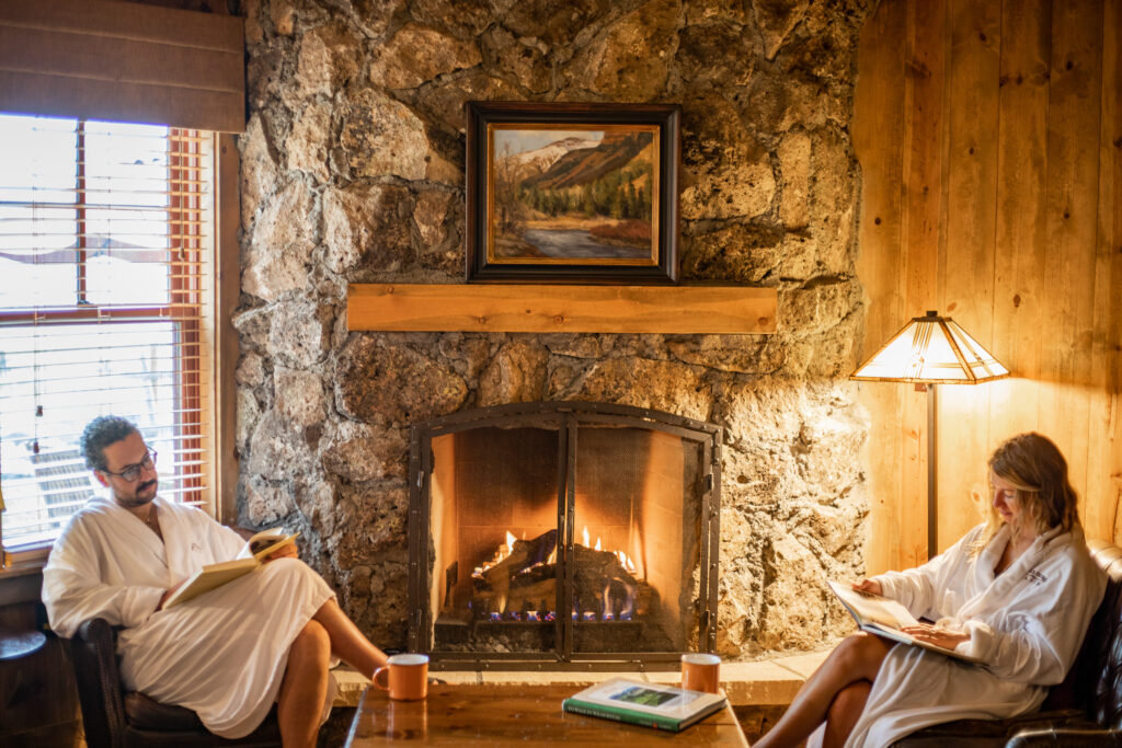 A man and woman dressed in robes sit by a lit fireplace reading.